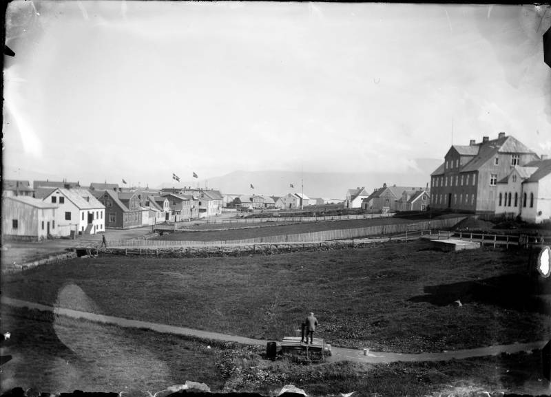 1902, Lækjargata í Reykjavík við upphaf nýrrar aldar, dönsk flögg blakta víða á húsþökum. Í forgrunni sækir aldurhniginn maður sér vatn úr Skálholtslind, vatnsbrunninum við Lækjargötu þar sem Mæðragarðurinn er nú. Þótt mikil breyting hafi orðið á götumyndinni eru nokkur hús sem standa enn. Lækurinn sem hér rennur úr Tjörninni og til hafs var settur í stokk árið 1911-1912. Soffía Claessen © Ljósmyndasafn Reykjavíkur